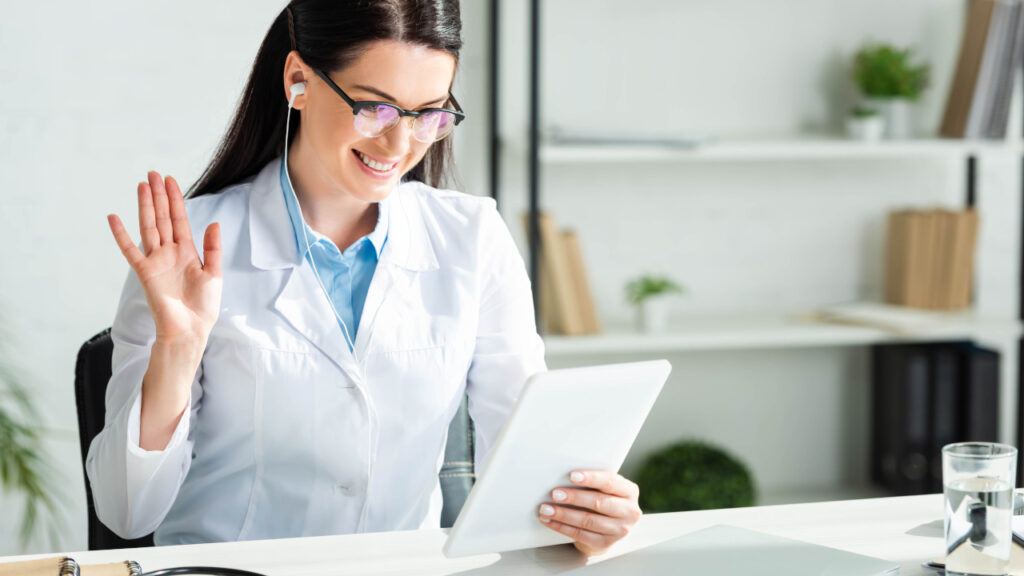 Médica do sexo feminino acenando para a tela de um tablet enquanto o segura com a mão esquerda.